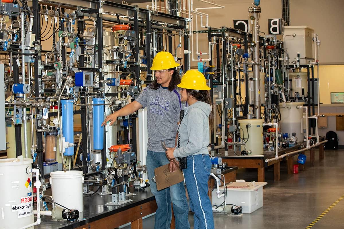 Two San Juan students in yellow hard hats look at a clipboard with machinery in the background